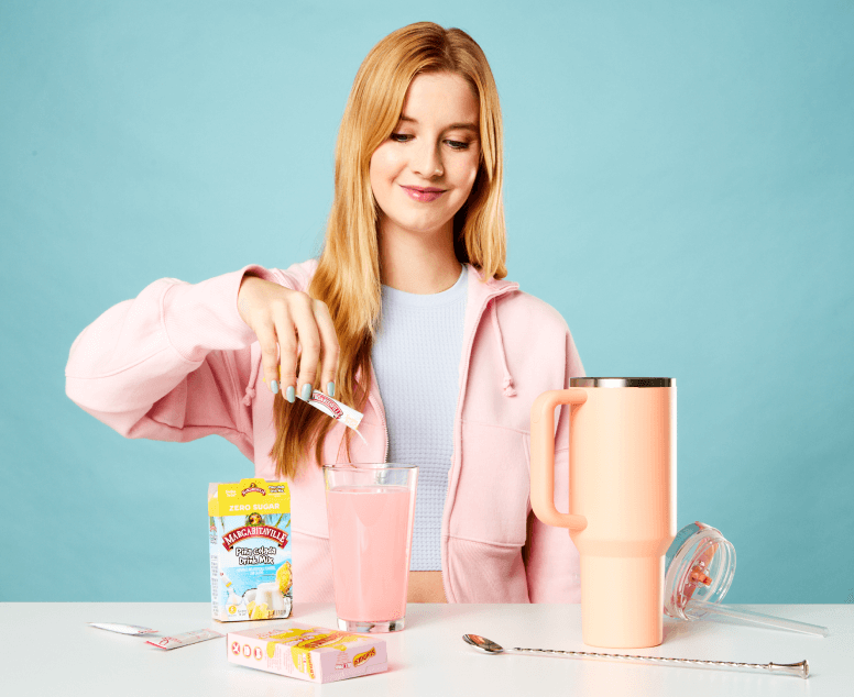 In the image, a woman with blonde hair is pouring a pink powder from a sachet into a glass of water. She's wearing a pink zip-up hoodie and a light blue shirt, coordinating with the pastel theme. o her right is a tall peach-colored tumbler with a lid off to the side, suggesting she's preparing a flavored drink. A straw and an open sachet are also visible on the table, set against a teal background, creating a fresh and bright setting.