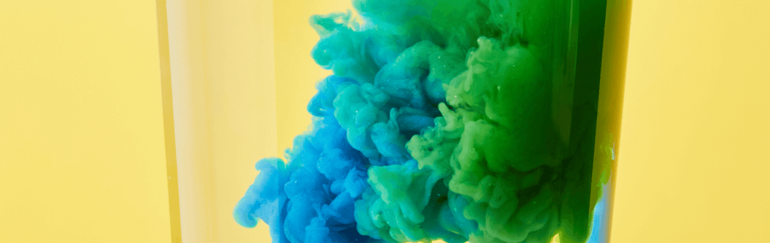 Vibrant blue and green water-soluble powder colors dispersing dynamically in a glass of water against a bright yellow background, illustrating the instant and lively blend of 'Singles to Go' drink mix.