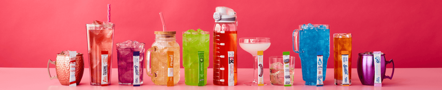 This image features a colorful display of various drinks in different types of containers set against a pink background. From left to right, there's a copper mug with a packet of cherry flavored drink mix, a tall glass with a straw holding a clear red beverage next to a Welch's packet